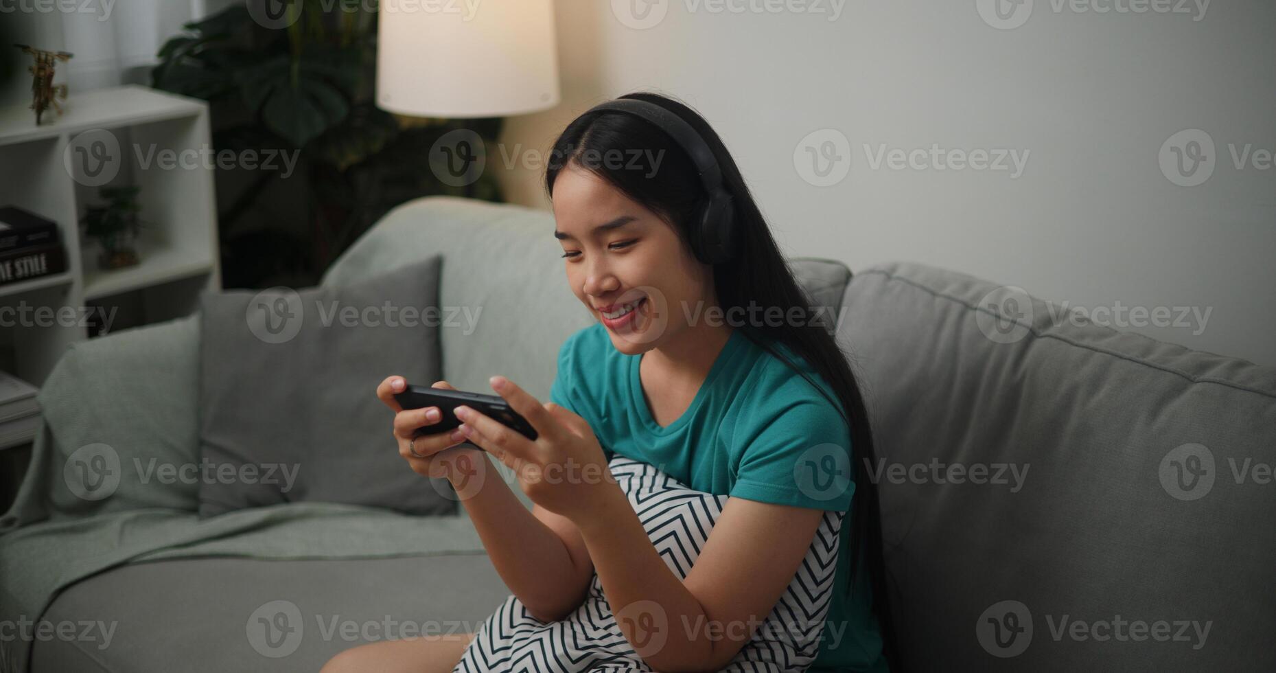Portrait of Young Asian woman wearing glasses and headphones enjoys playing online esport games on smartphone sitting on sofa in the living room at home,Gamer lifestyle concept. photo