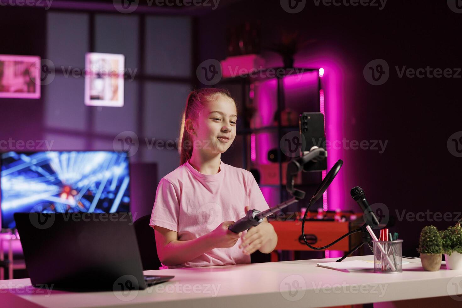 Kid shooting product unboxing for streaming channel on online platforms using cellphone on selfie stick. Cute girl warmly welcoming fans, filming reviews with mobile phone camera photo
