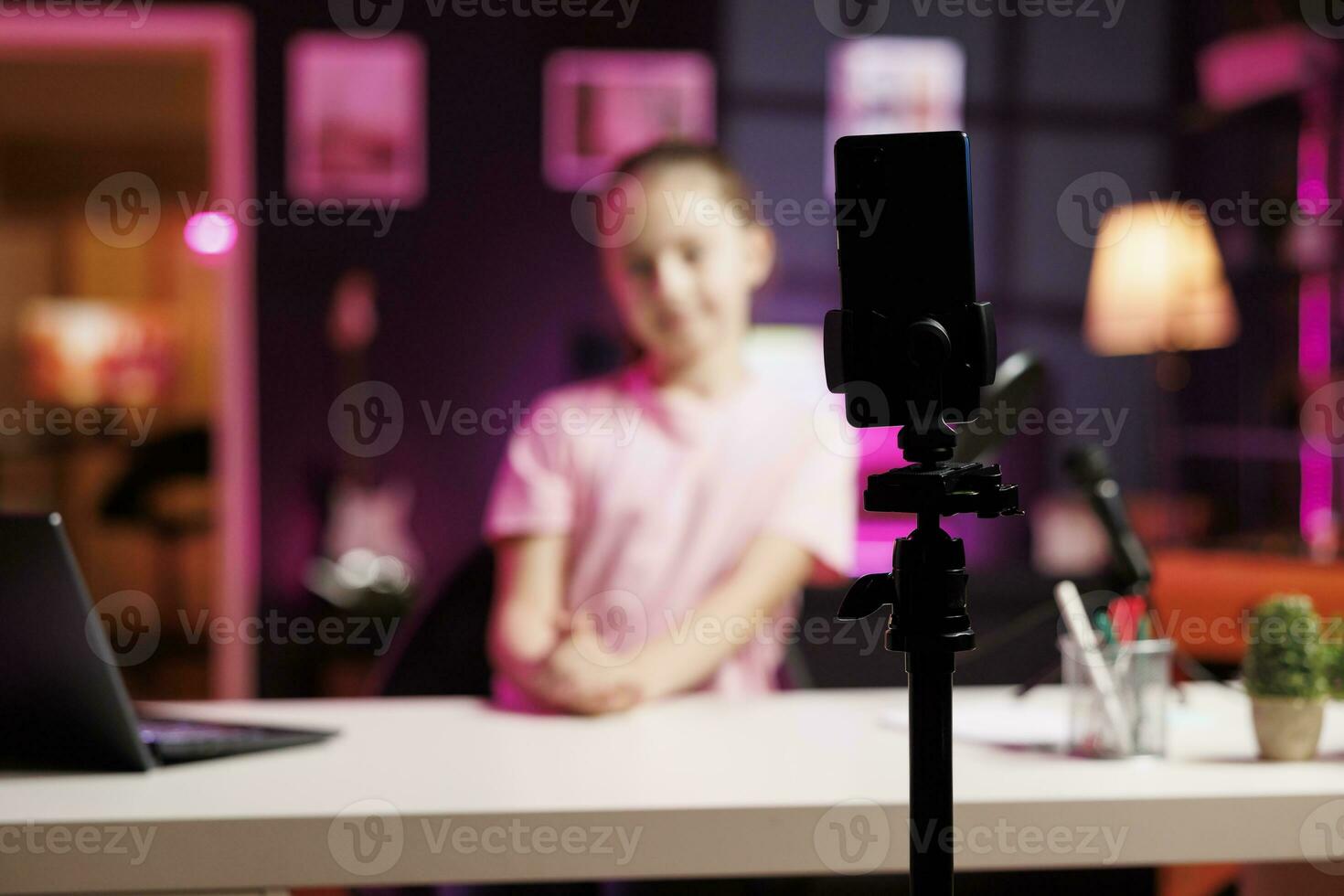 Young child in blurry background doing livestream using smartphone on professional recording tripod. Cute kid does live broadcast using cellphone camera to talk with online audience, focus on phone photo