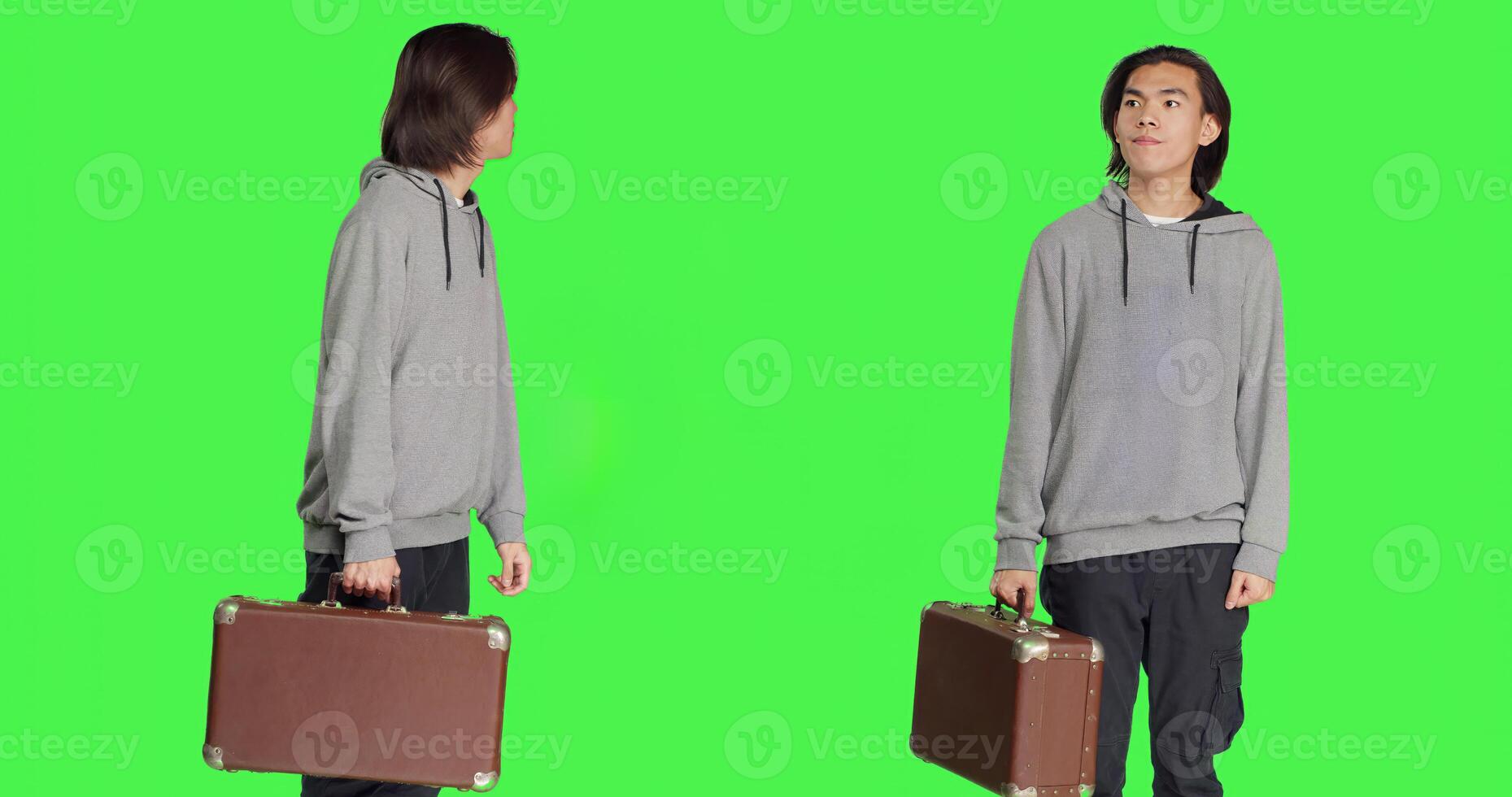 Young person carries vintage briefcase over greenscreen template in studio, waiting impatiently for something. Asian guy holds big suitcase while he checks time on wrist watch. photo