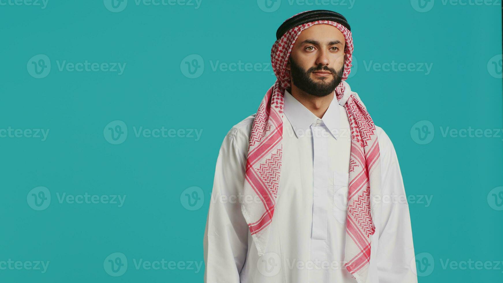 Middle eastern man in traditional attire smiling and posing with confidence, representing arabic cultural identity with checkered print headscarf and white robe. Young cheerful arab person. photo