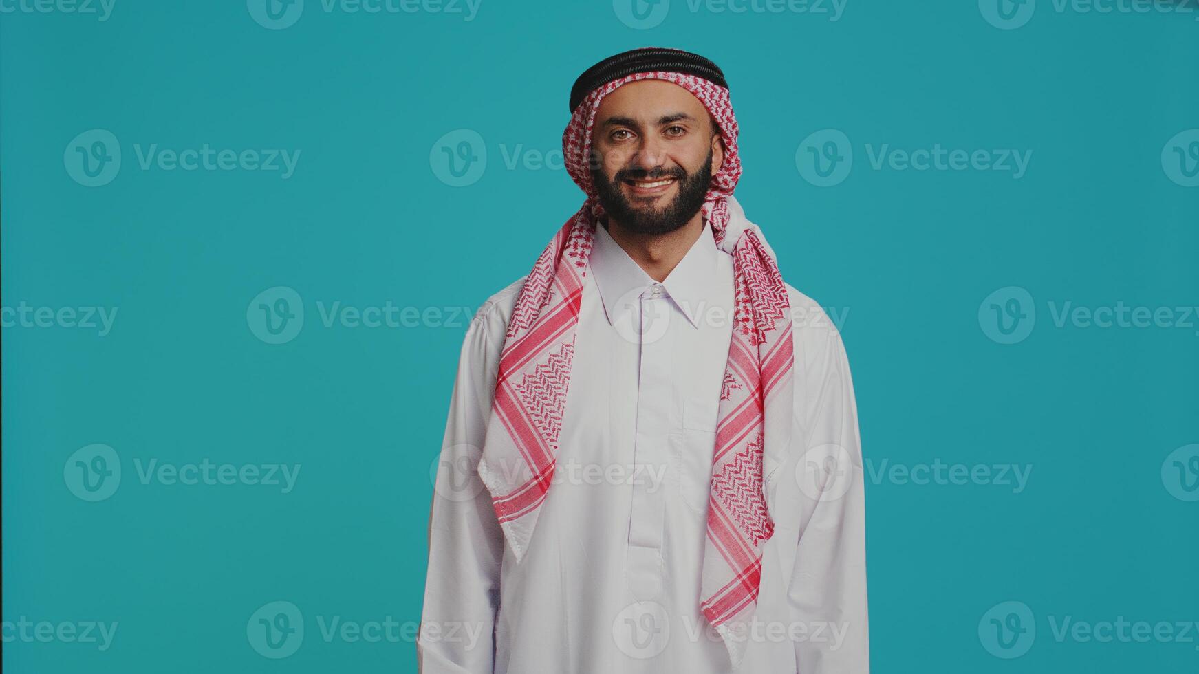 Middle eastern adult in religious attire smiling and posing proudly, representing muslim cultural identity with red and white print headscarf and robe. Young confident islamic person. photo