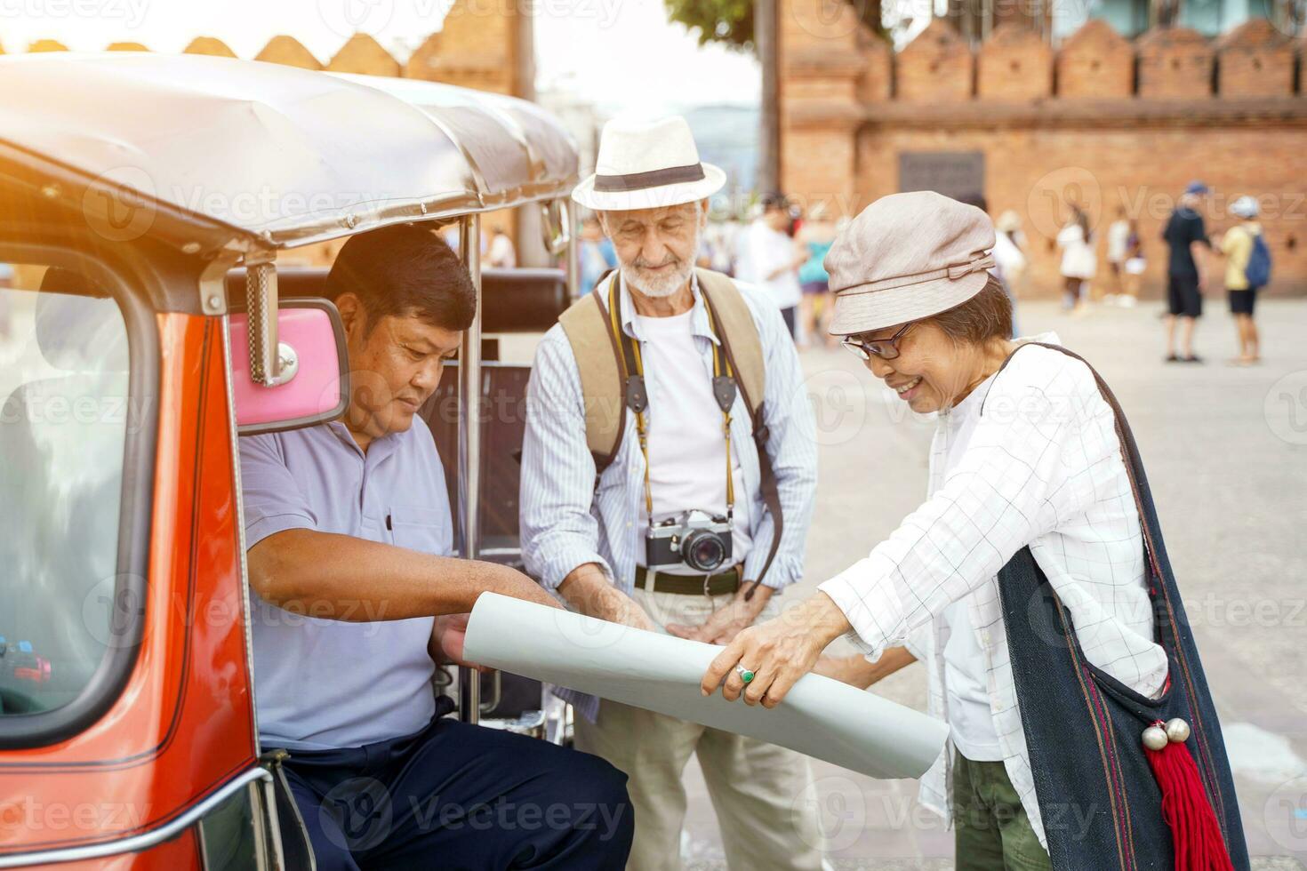 American senior tourist man with his tour guide ask the Tuktuk Thailand taxi driver for them destination on the map with sun bright and blurred of city gate background. Senior tourist concept photo