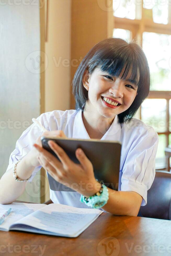 Closeup and front view of happiness cheerful Thai girl student holding a tablet with studying in the classroom on sun flare background. Network education concept. photo