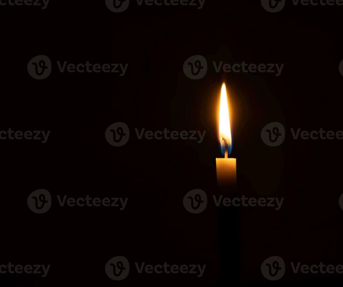 A single burning candle flame or light glowing on small orange candle on black or dark background on table in church for Christmas, funeral or memorial service with copy space photo