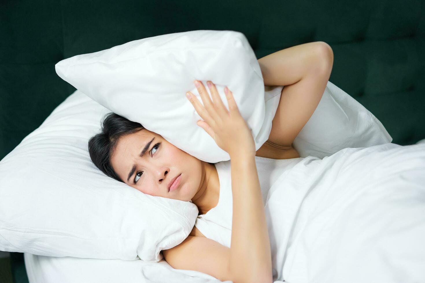 Insomnia. Asian girl shuts her ears with pillow as partner snoring. Woman lying in bed, bothered by noise at night, cannot sleep photo