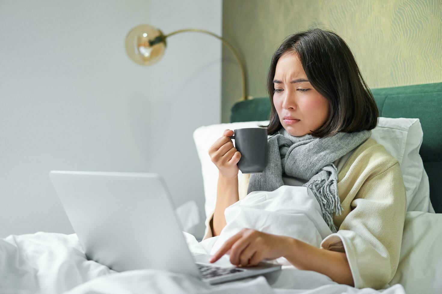 asiático niña mirando a computadora portátil, sensación enfermo, atrapando frío, Bebiendo caliente té para dolorido garganta, ceñudo como mirando a computadora pantalla foto