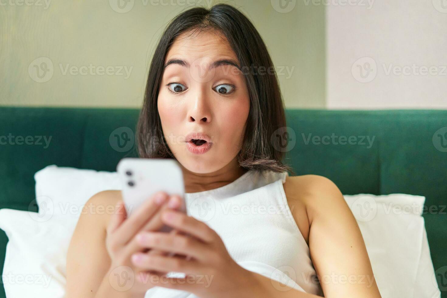 Cute asian girl holding smartphone, looking surprised at mobile phone screen, reading message with amazed face expression photo