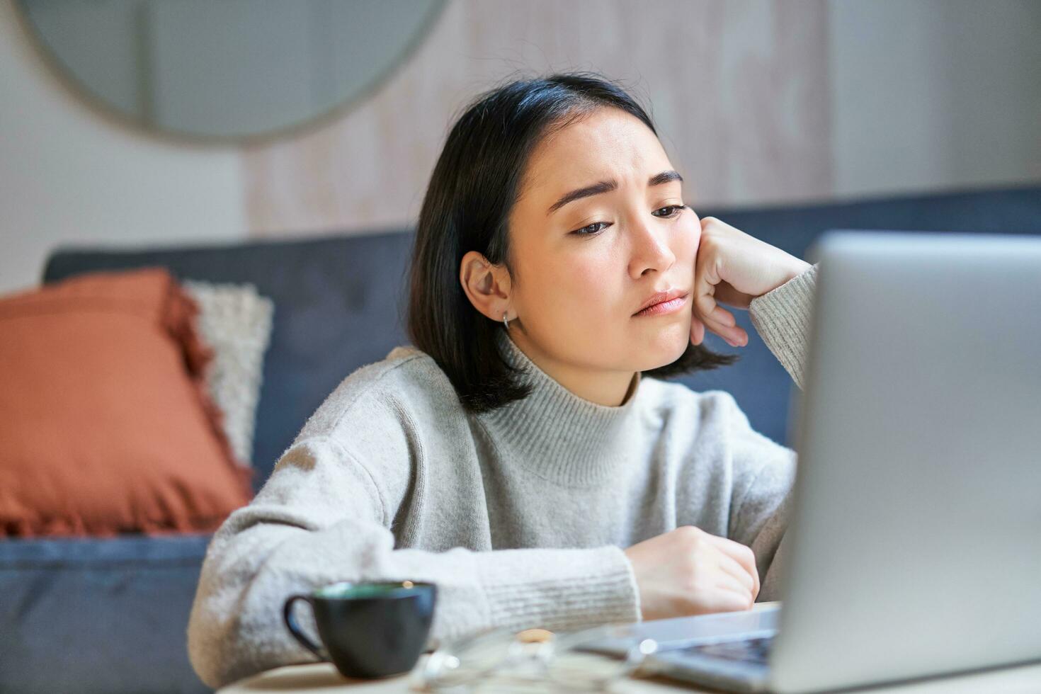 retrato de coreano niña se sienta aburrido, estudiante mira melancólico a computadora portátil, sentado a hogar y expresando aburrimiento foto