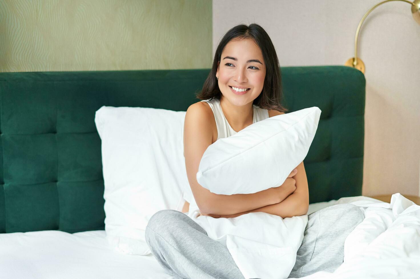 Romantic smiling asian girl lying in her bed, hugging pillow and gazing dreamy, lazy mornings in bedroom photo