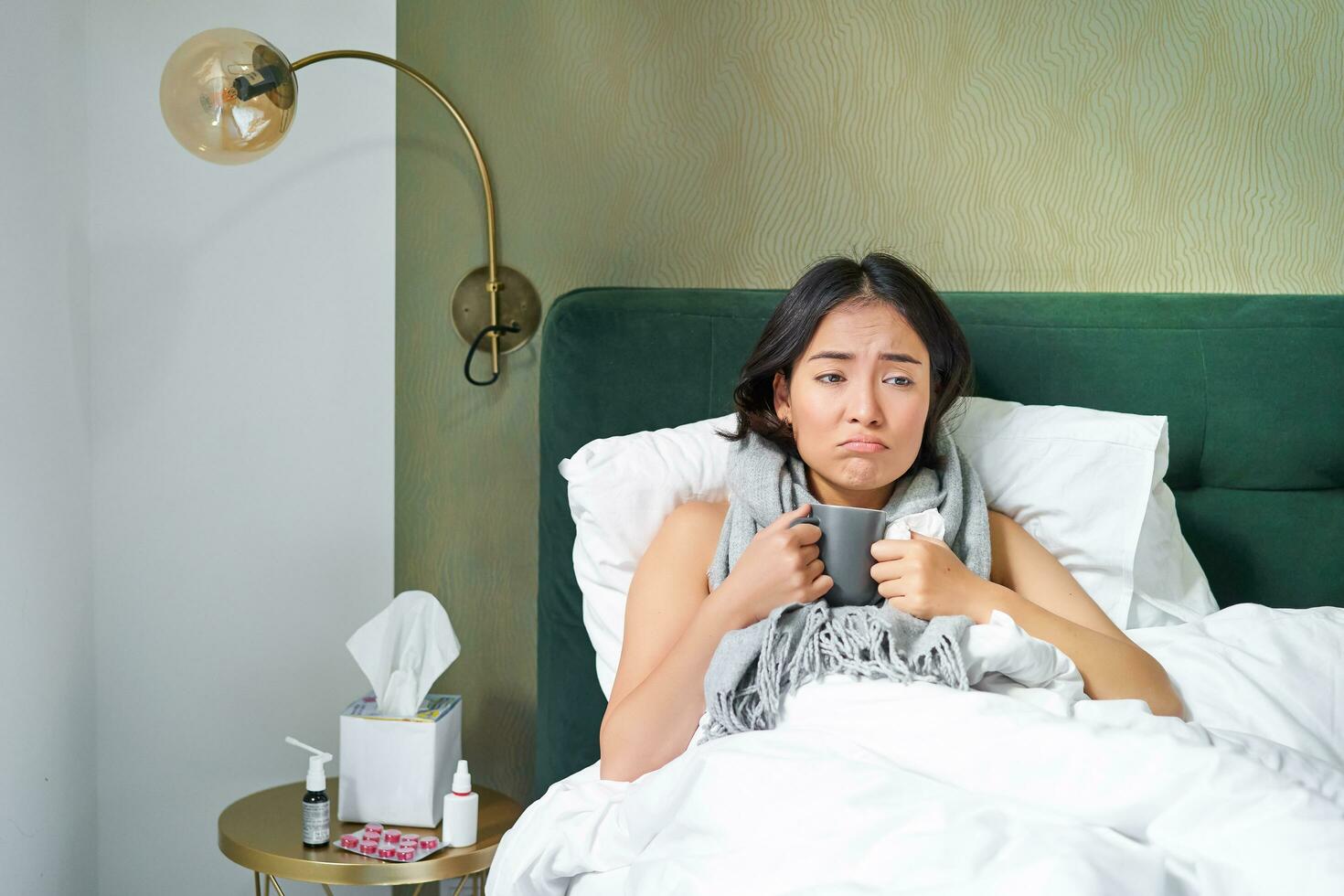 Sick sad asian girl lying in her bed with flu, catch a cold, taking pills and drinking hot tea, feeling upset photo