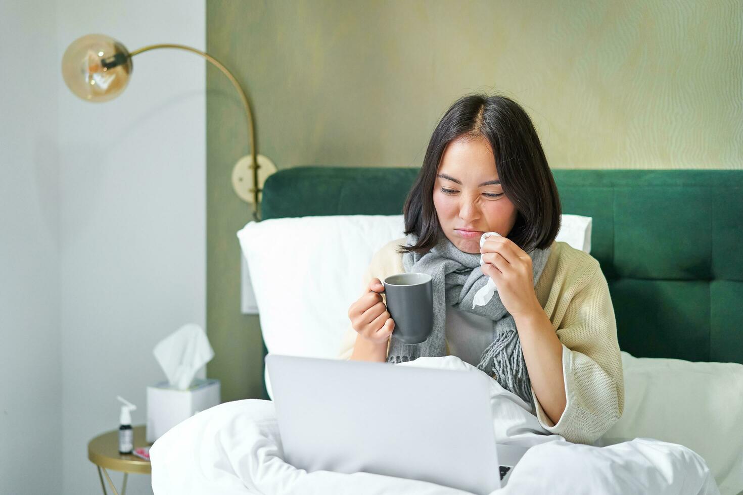 Hardworking asian girl staying in bed with cold, catching flu but working from home with laptop, studying while feeling sick photo