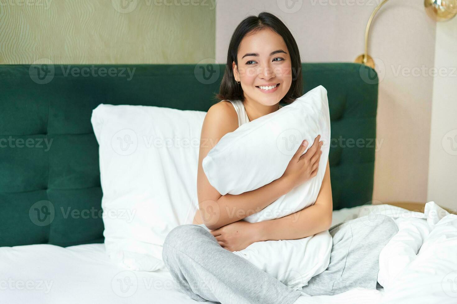Romantic smiling asian girl lying in her bed, hugging pillow and gazing dreamy, lazy mornings in bedroom photo