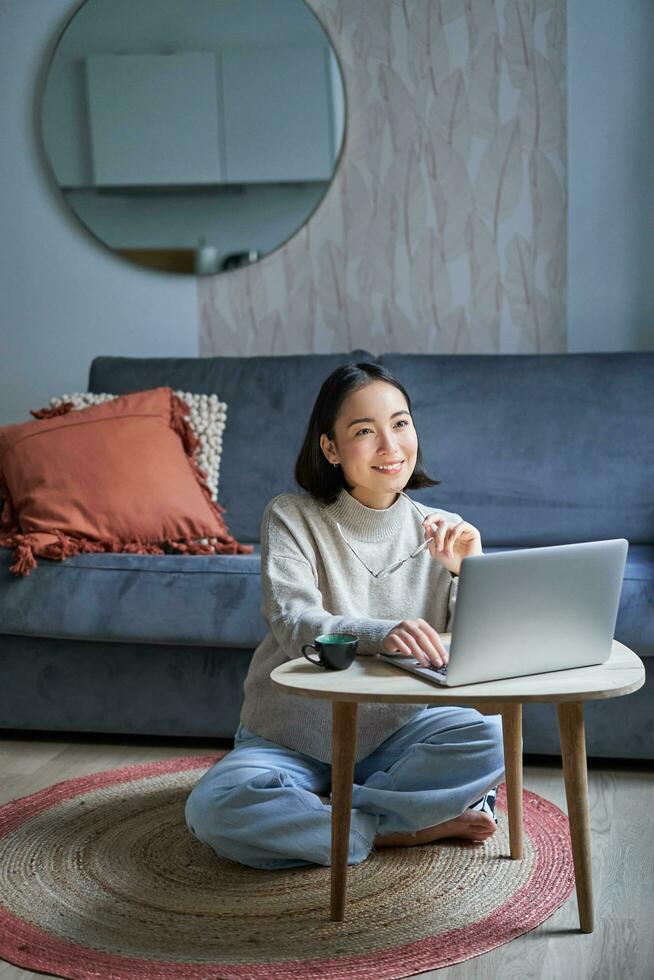 Vertical shot of korean working woman, sitting on floor at home with laptop, studying, using computer on remote, e-learning concept photo