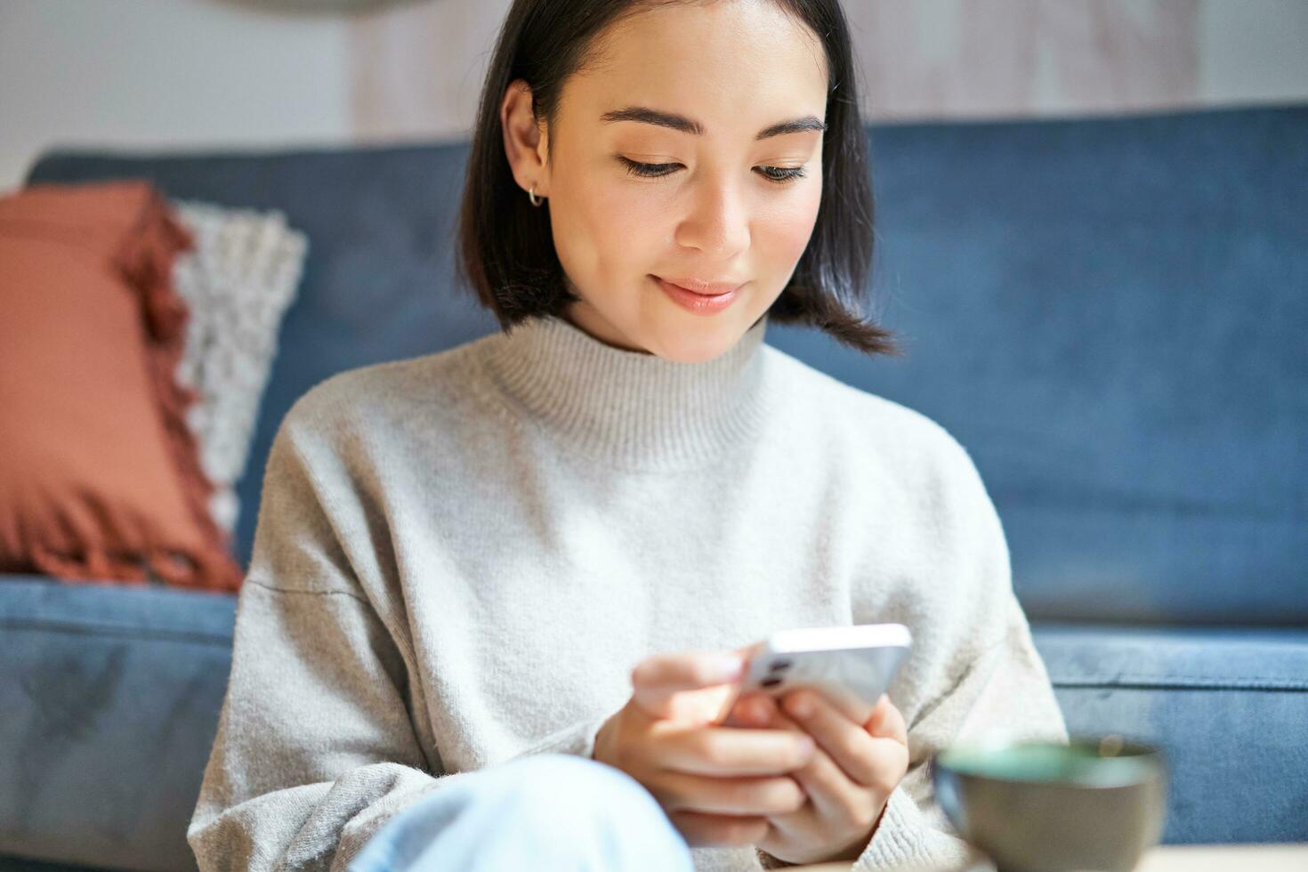 Portrait of smiling asian woman sits at home, uses smartphone app, orders delivery, messages someone on mobile phone application photo