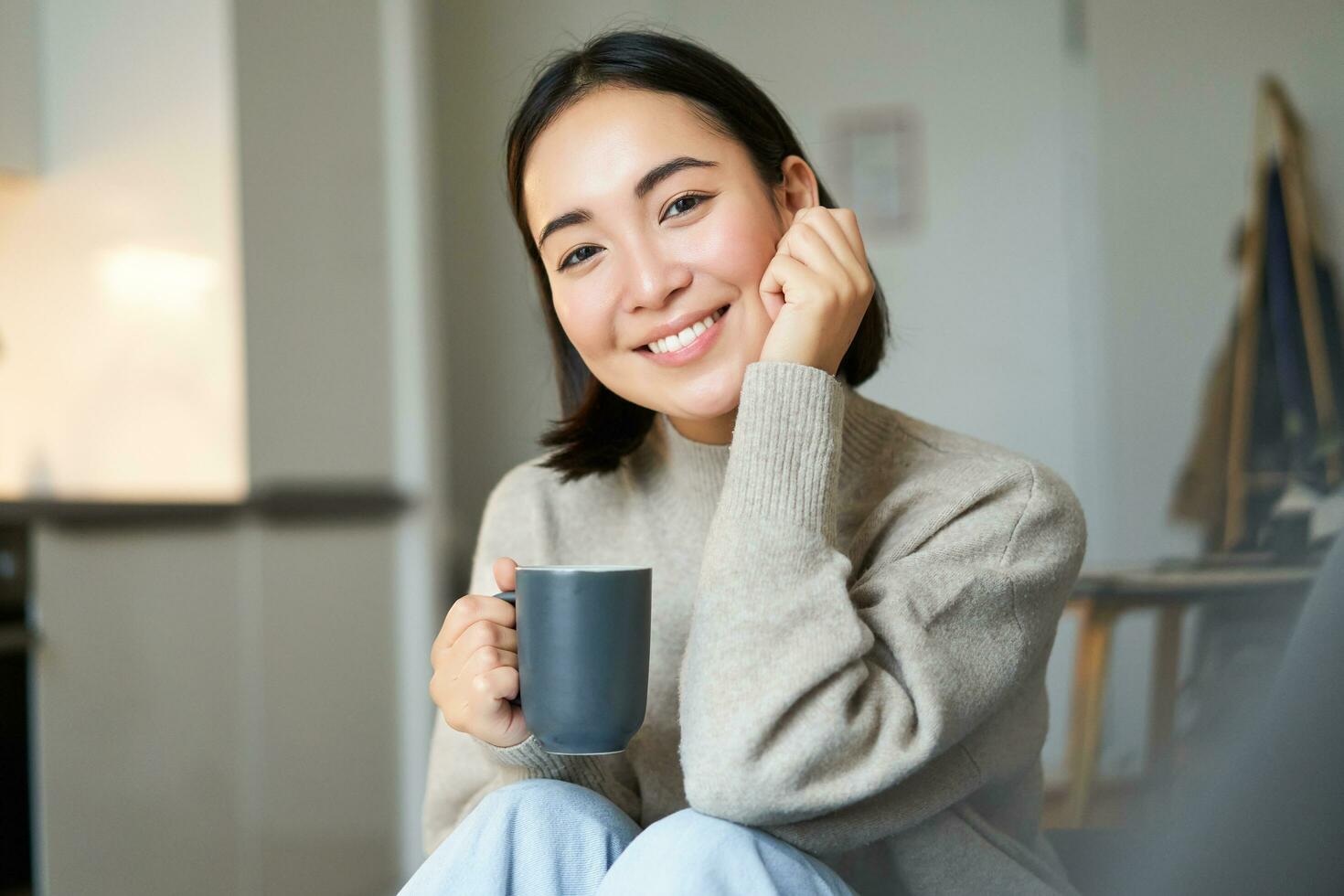 Smiling asian woman sitting at home with cup of coffee, relaxing and feeling warmth, looking outside window, resting on sofa in living room photo