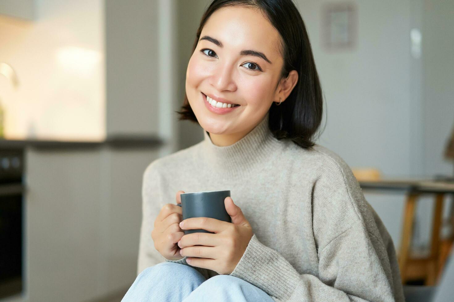 retrato de sonriente asiático niña con taza de café, Bebiendo caliente té y consiguiendo acogedor a hogar. calentamiento arriba con bebida foto