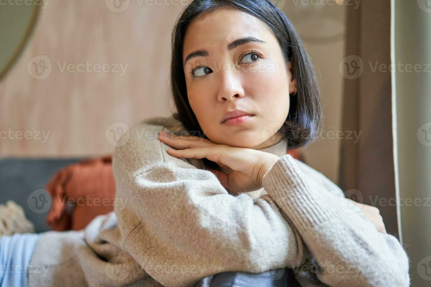 Women and wellbeing concept. Portrait of thoughtful asian woman sitting on sofa with pillow, looking aside with thinking concentrated face photo