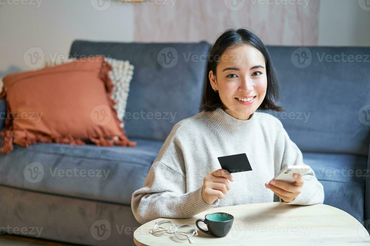 Smiling cute asian woman using credit card and smartphone, paying bills online, holding mobile phone, looking at camera photo