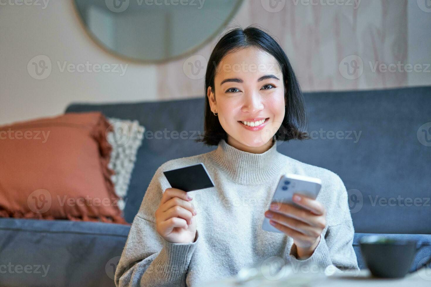 Portrait of smiling korean woman using credit card and smartphone app, paying for purchase, order online delivery photo