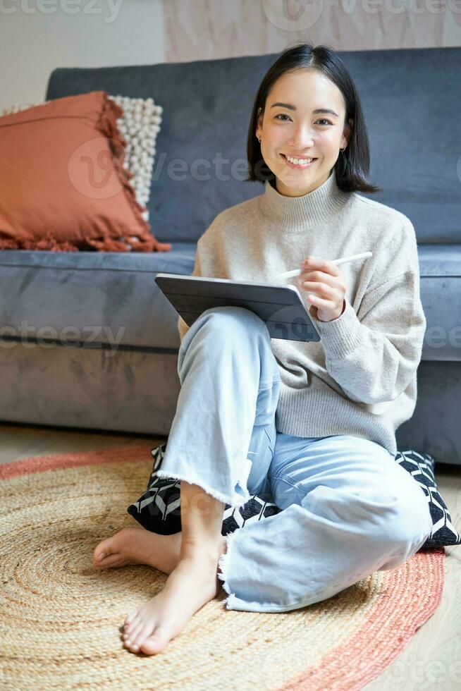 Vertical shot of cute asian girl in cozy home, drawing on her digital tablet with pen, designer working on project photo