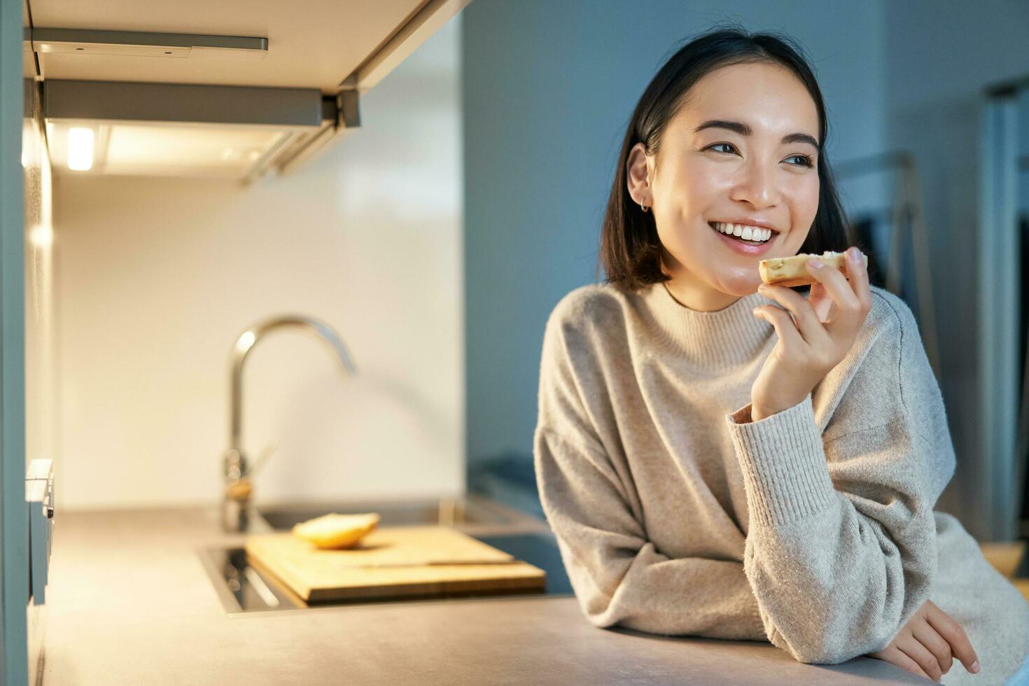 retrato de sonriente joven contento mujer quedarse a hogar, en pie en cocina y comiendo tostada, mirando aparte foto