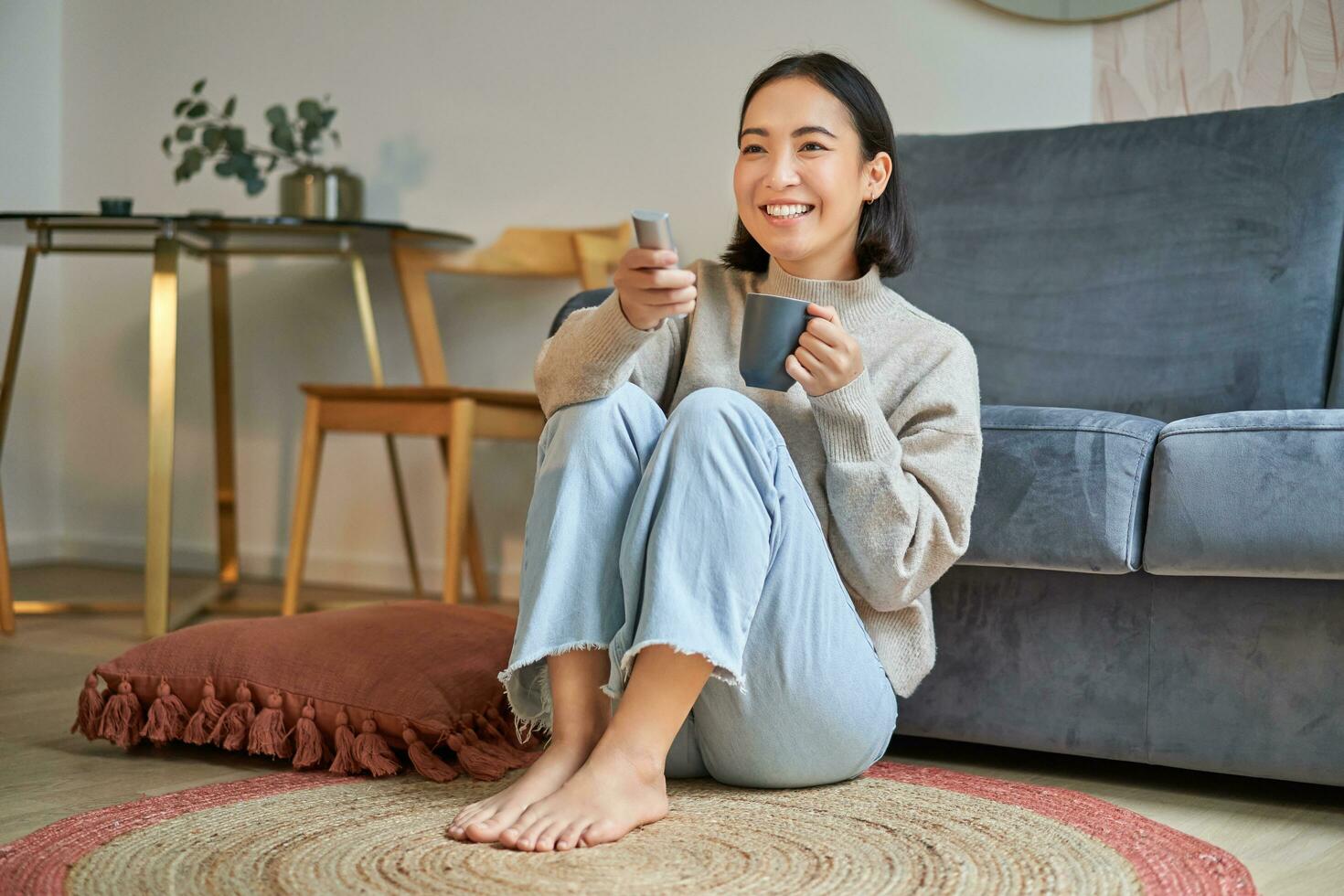 Portrait of girl watching television at home, sits on floor near sofa, holds remote and changes channels photo