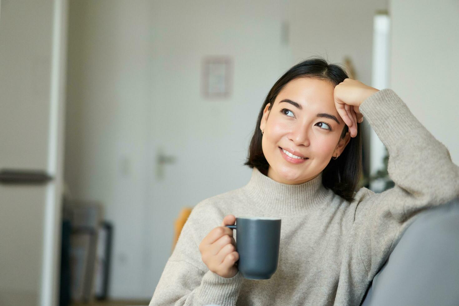 Smiling asian woman sitting on sofa with her mug, drinking coffee at home and relaxing after work, looking calm and cozy photo