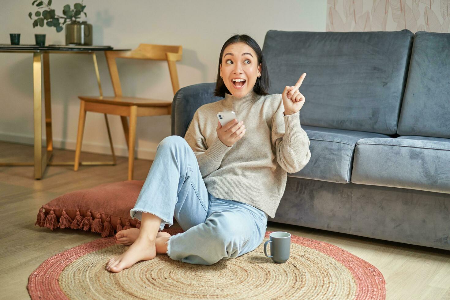 Portrait of modern young korean woman sitting on floor, pointing finger at copy space, showing banner or advertisement, holding smartphone as using app photo