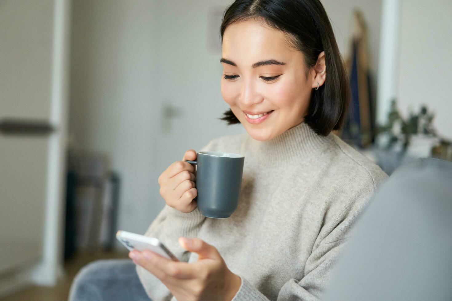 Portrait of smiling asian girl checking her news feed on smartphone and drinking coffee, sitting on sofa at home, browsing on mobile phone, reading photo