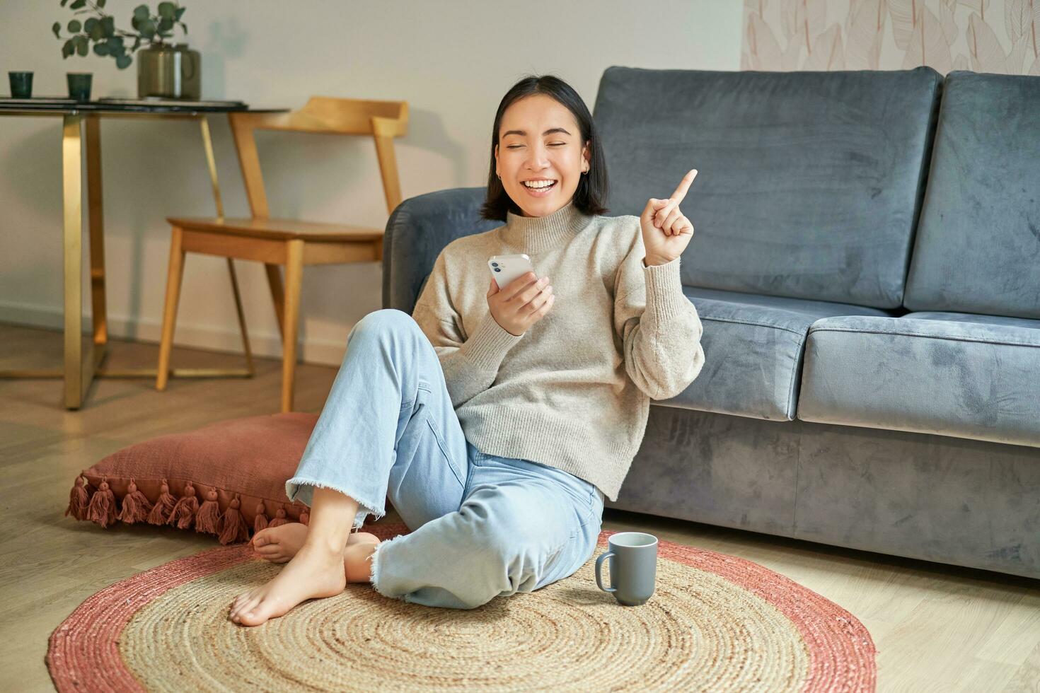 Portrait of modern young korean woman sitting on floor, pointing finger at copy space, showing banner or advertisement, holding smartphone as using app photo
