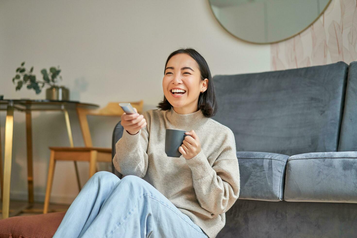 Portrait of beautiful asian girl sitting at her home and watching tv, holding remote, smiling and laughing, feeling comfort and warmth at her apartment photo