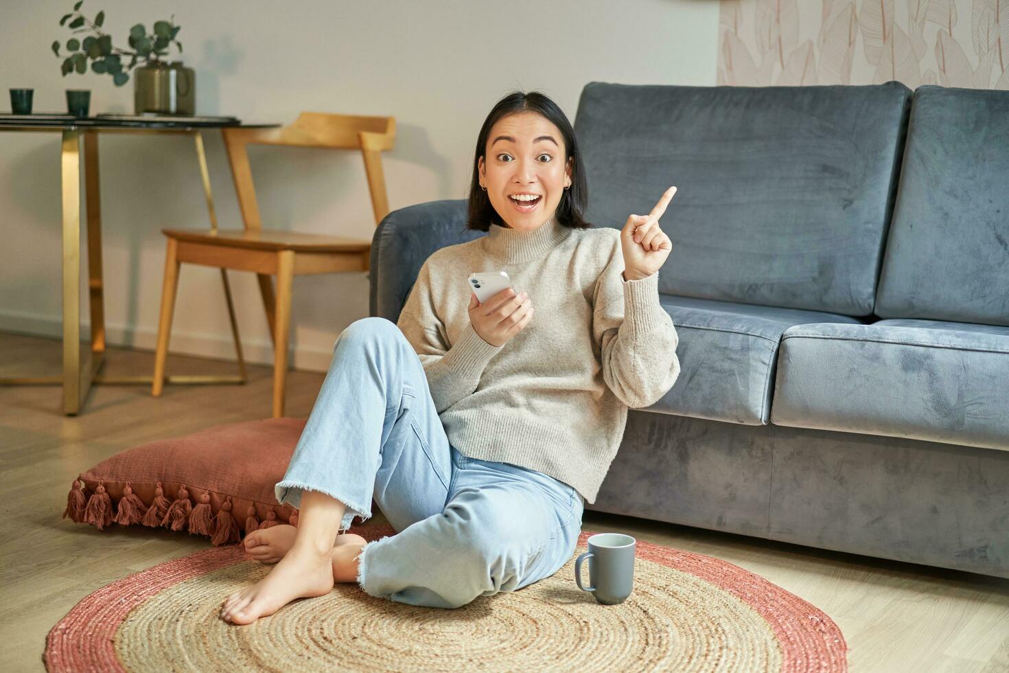 Smiling asian girl sits on floor in stylish living room, pointing finger at advertisement, showing promo banner, holding mobile phone in hand photo