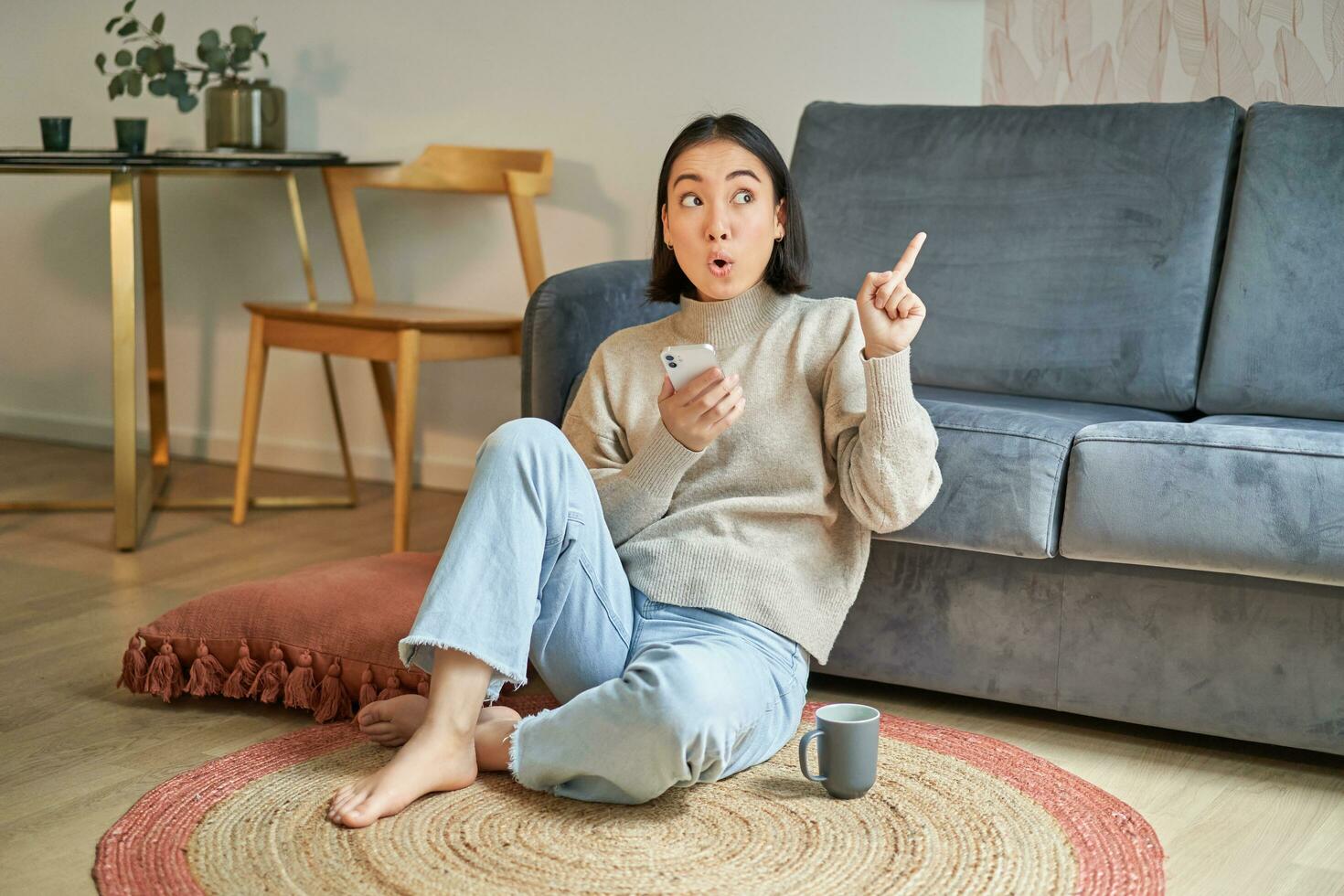 Portrait of modern young korean woman sitting on floor, pointing finger at copy space, showing banner or advertisement, holding smartphone as using app photo