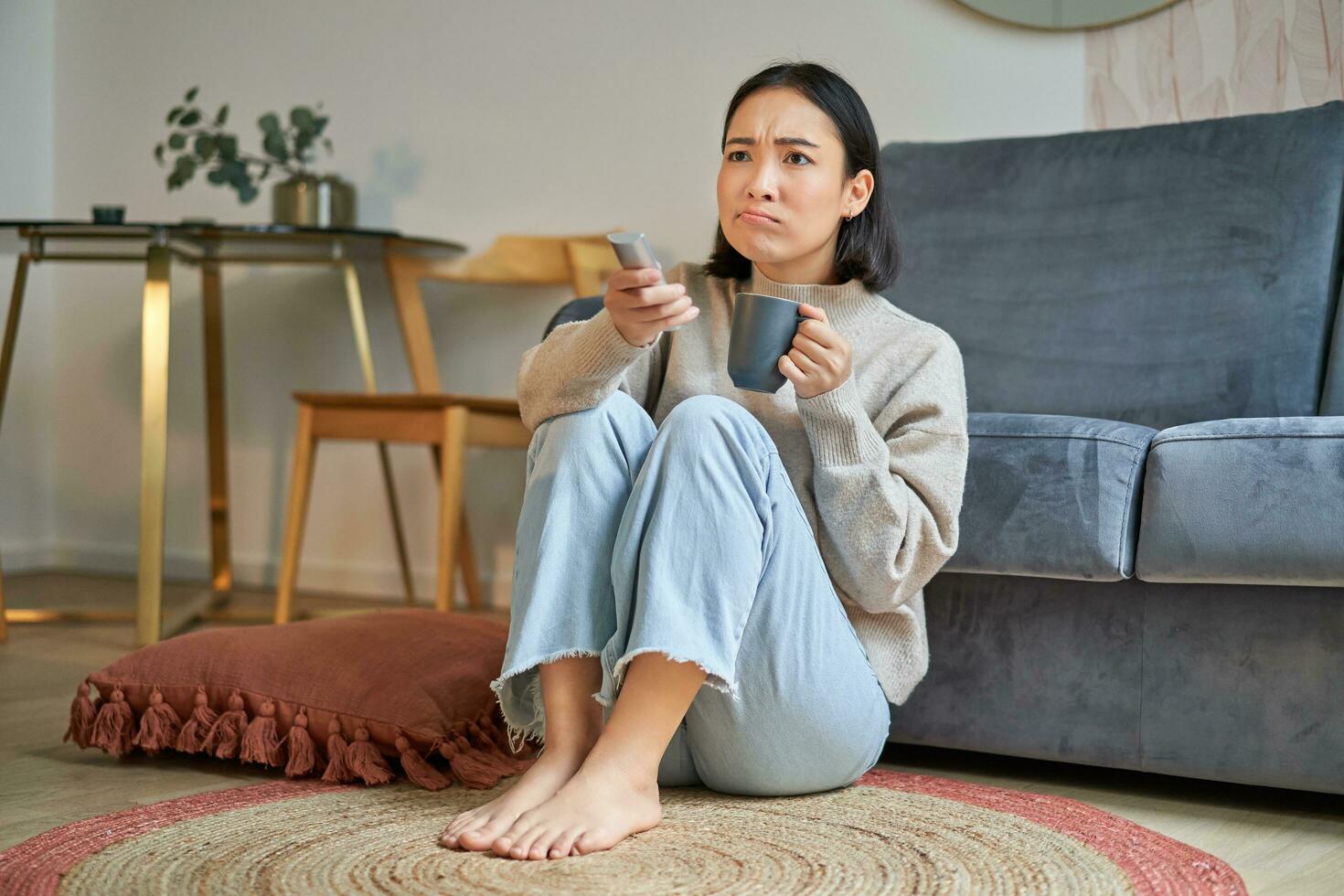 Portrait of girl watching television at home, sits on floor near sofa, holds remote and changes channels photo