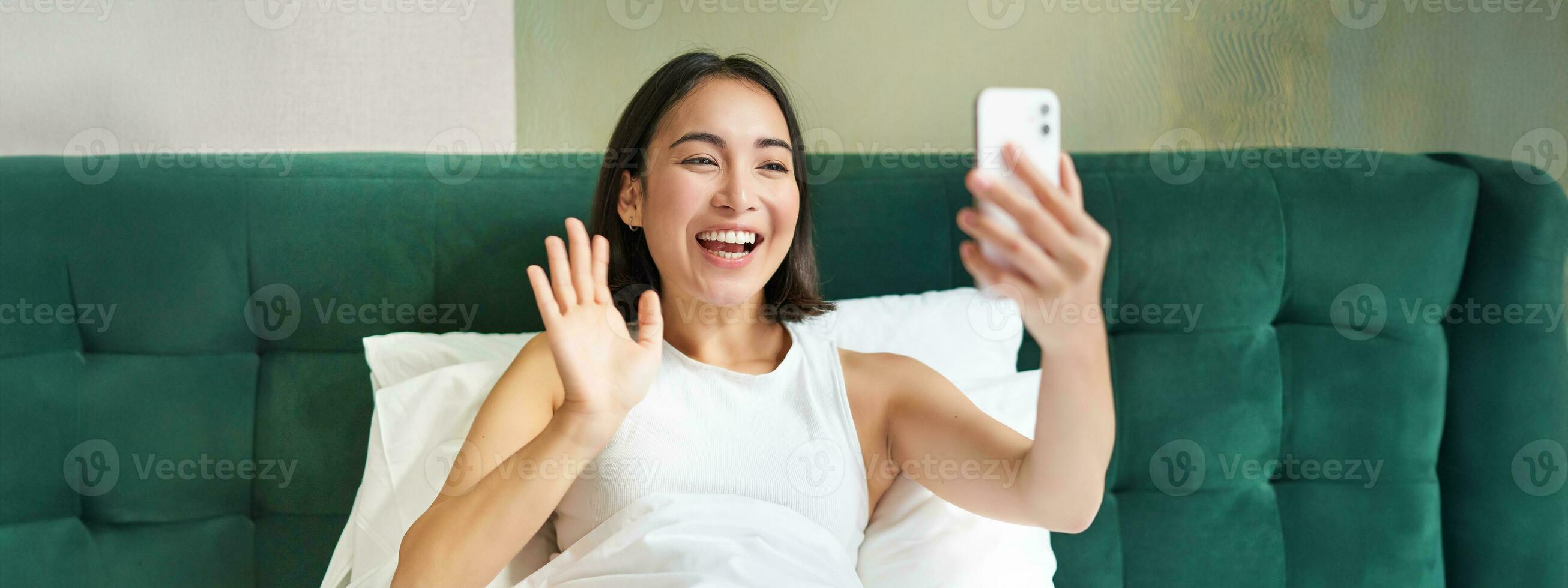 Portrait of smiling korean woman looking at smartphone, lying in bed and video chatting, waving hand at mobile phone camera, taking selfie, recording video photo