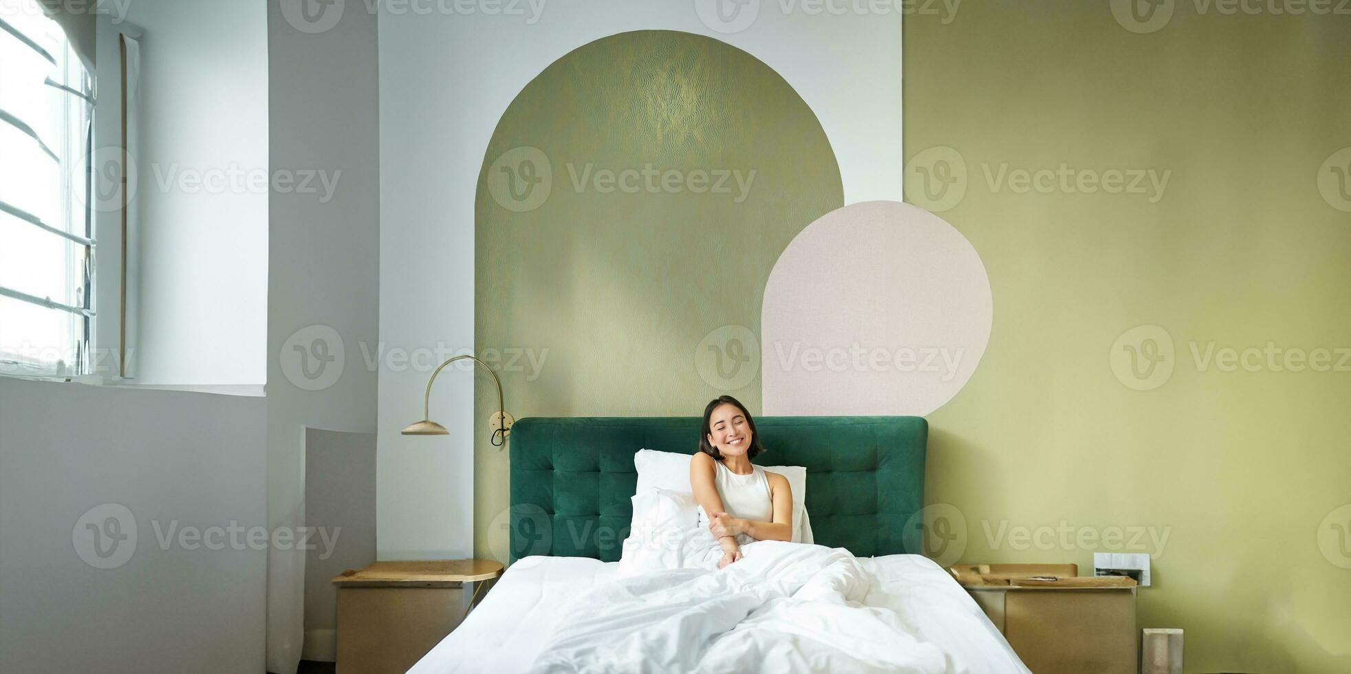 Vertical shot of beautiful asian girl lying in bedroom with sophisticated interrior, smiling and looking happy in morning, spending time in bed photo