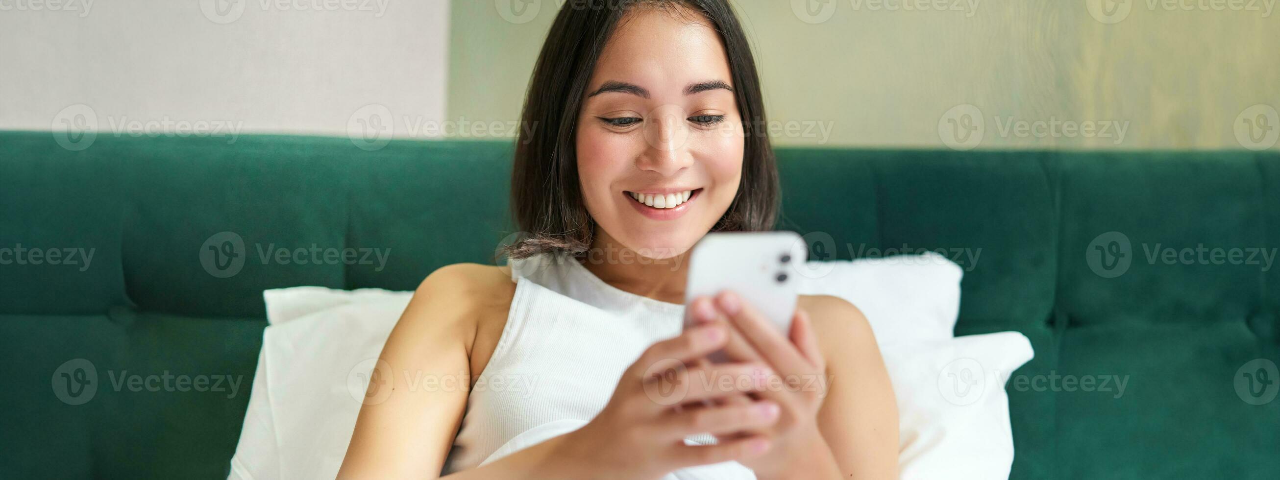 Portrait of smiling asian woman lying in bed, looking at smartphone, using mobile phone with happy face, relaxing on lazy morning photo