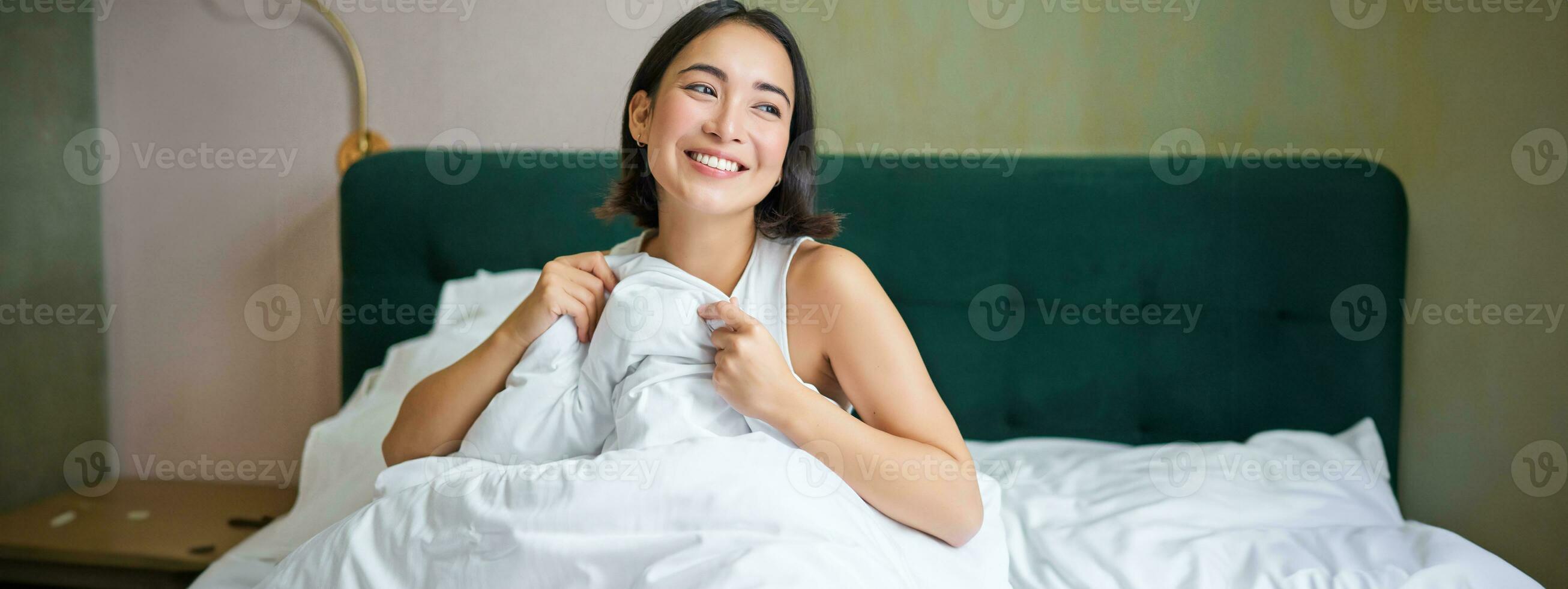 Close up of happy beautiful asian woman, waking up in bed and enjoys morning, looking outside window with sleepy smile on her face, hugging duvet photo