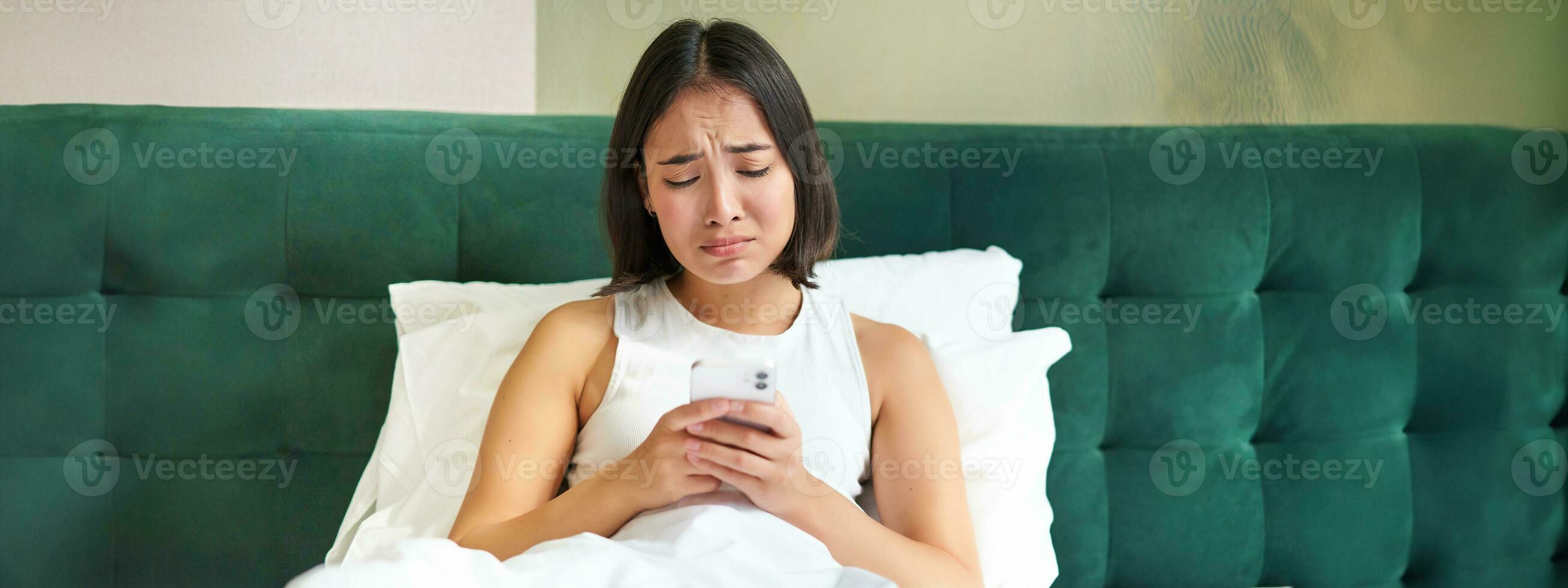 Disappointed asian girl, crying and whining, feeling upset, lying in bed on pillow, holding mobile phone, read upsetting bad news, waking up in awful mood photo