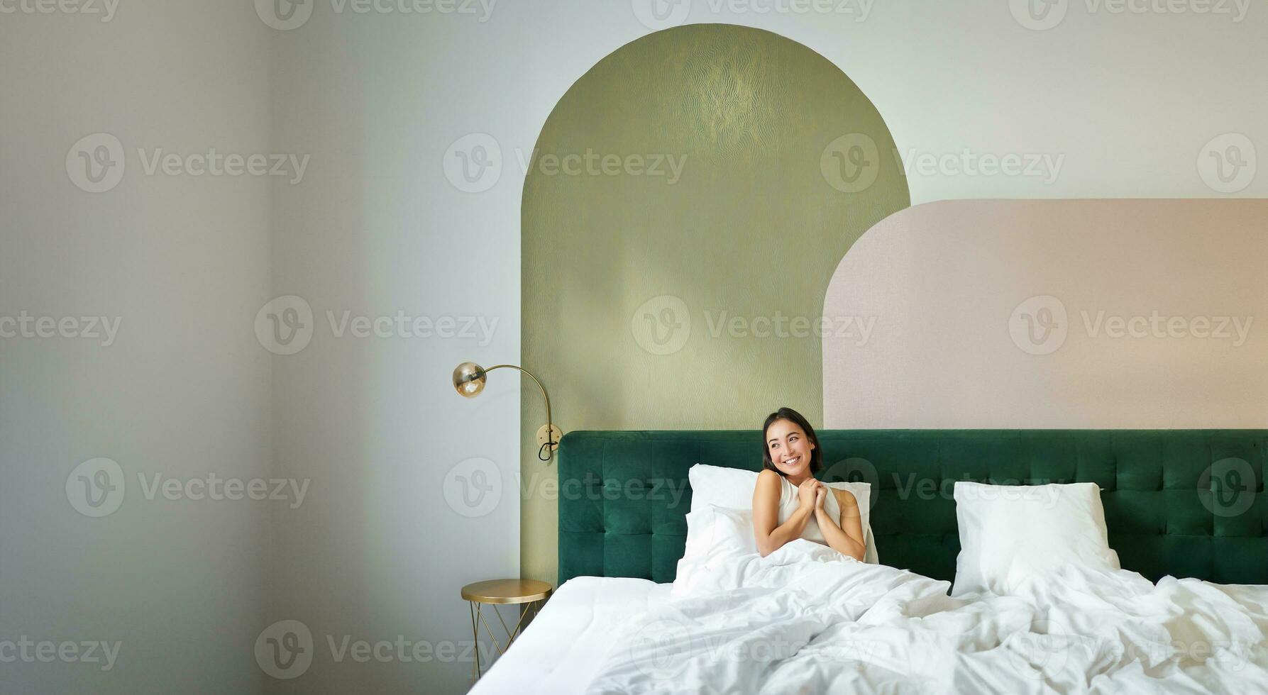 Vertical shot of beautiful asian girl lying in bedroom with sophisticated interrior, smiling and looking happy in morning, spending time in bed photo