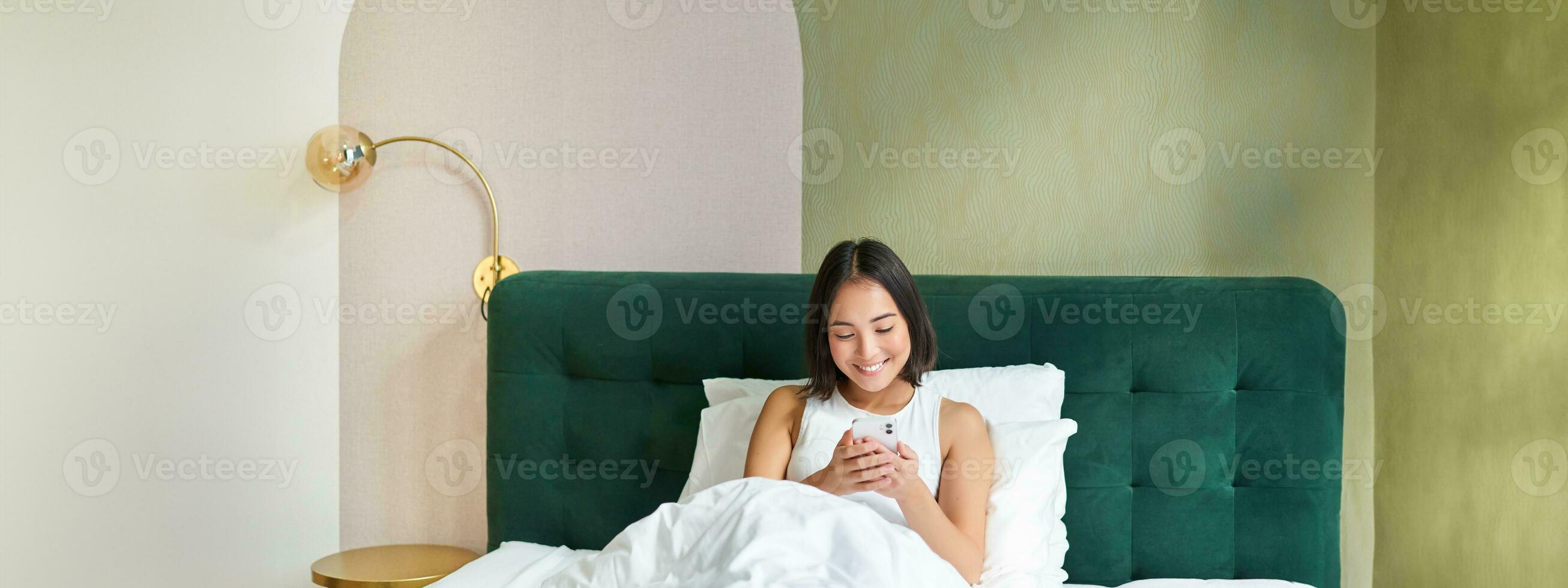 Portrait of smiling asian woman lying in bed, looking at smartphone, using mobile phone with happy face, relaxing on lazy morning photo