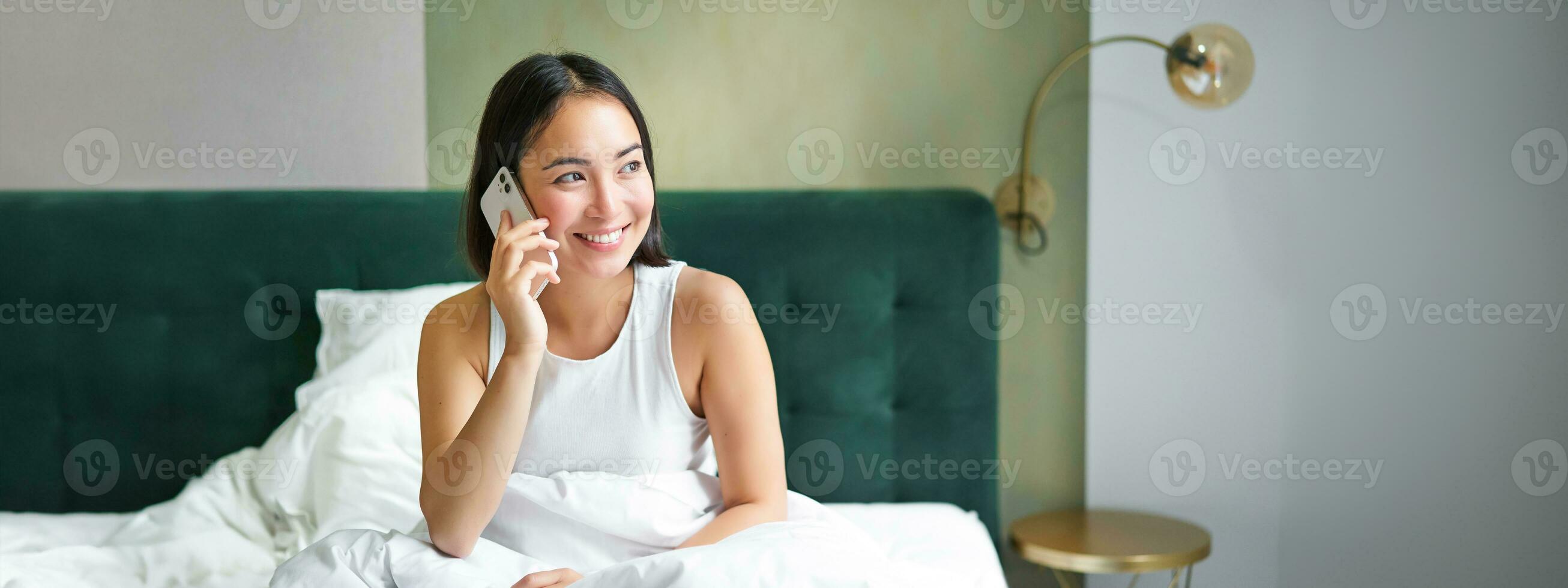 sonriente coreano niña en cama, negociaciones en móvil teléfono, haciendo un teléfono llamar, perezoso Mañana como asiático mujer pedidos entrega vía teléfono inteligente foto