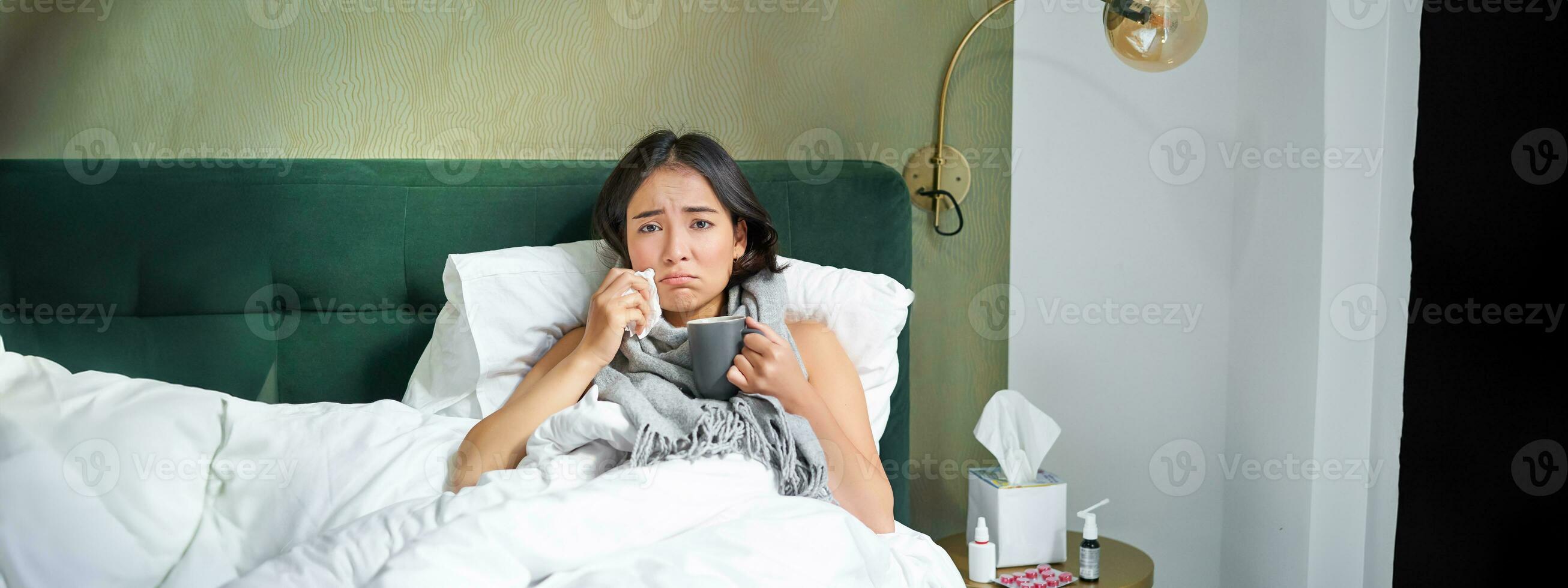 Health and people. Asian girl lying in bed, feeling sick, sneezing and drinking hot tea, catching cold, staying at home on leave photo