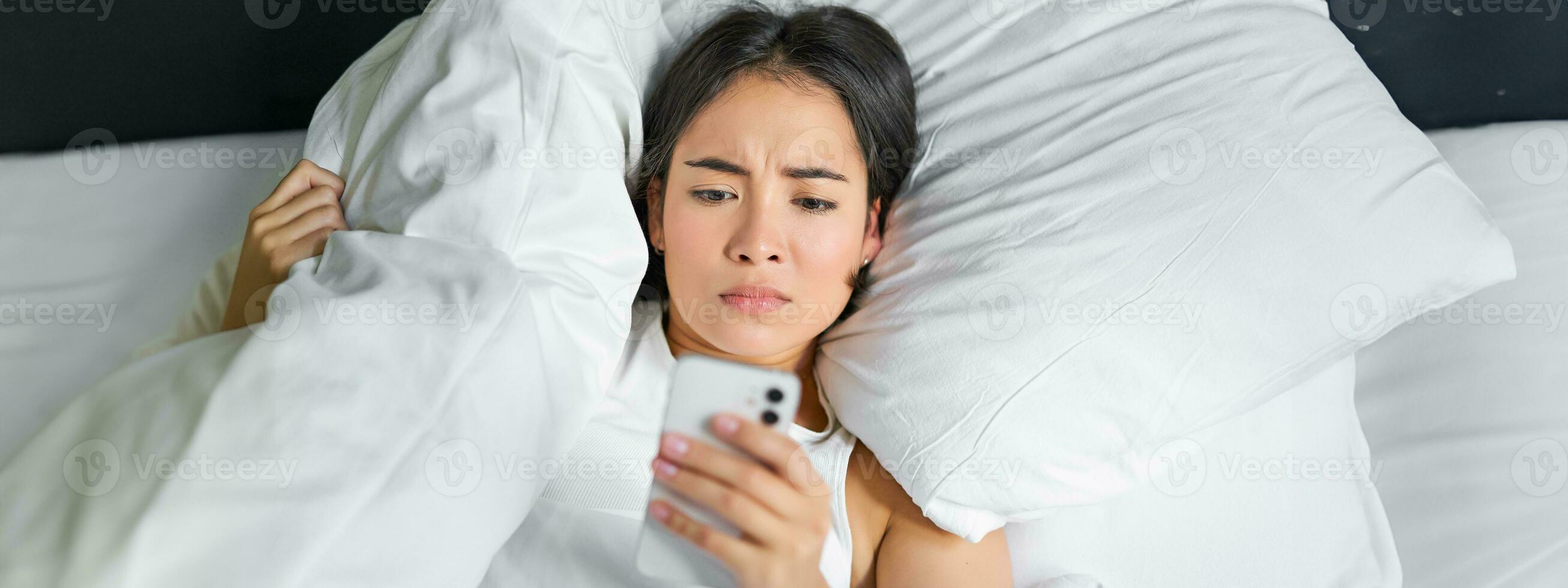 Close up portrait of asian girl lying in bed, looking at smartphone concerned, waking up late and staring at her alarm clock on mobile phone photo