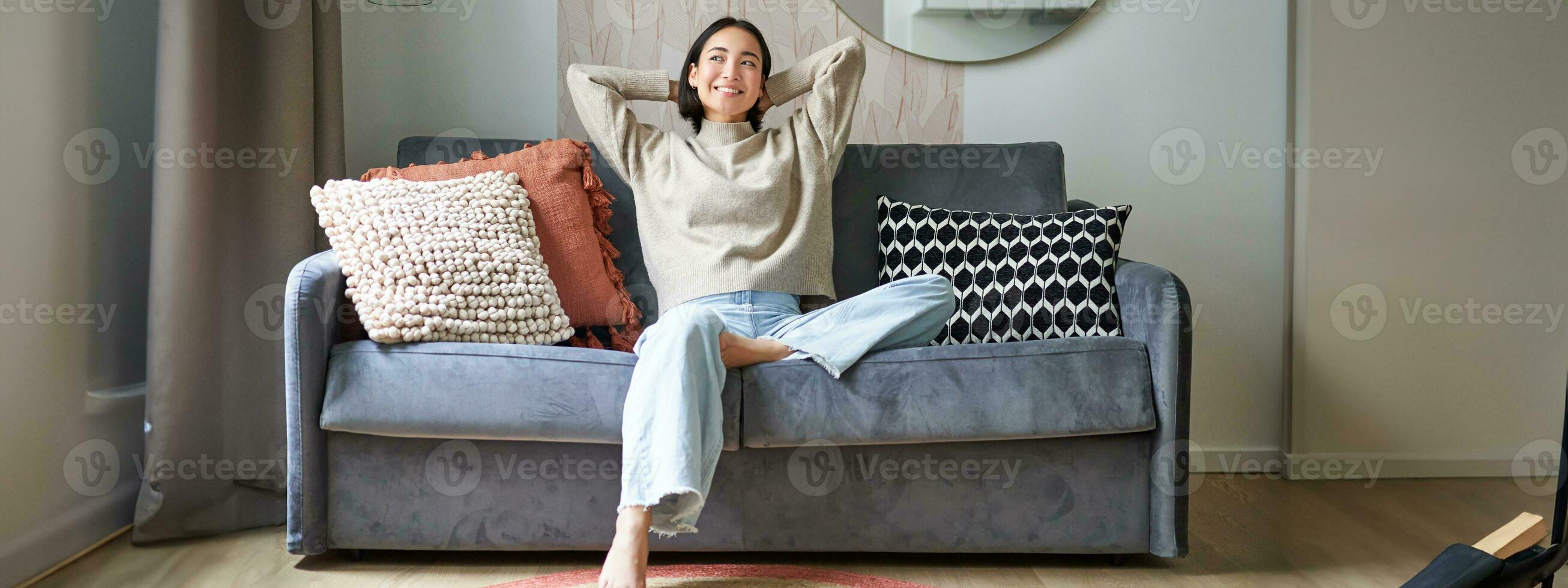 retrato de contento asiático mujer sensación perezoso, extensión en sofá y sonriente complacido, relajante a hogar, descansando desde trabajo foto