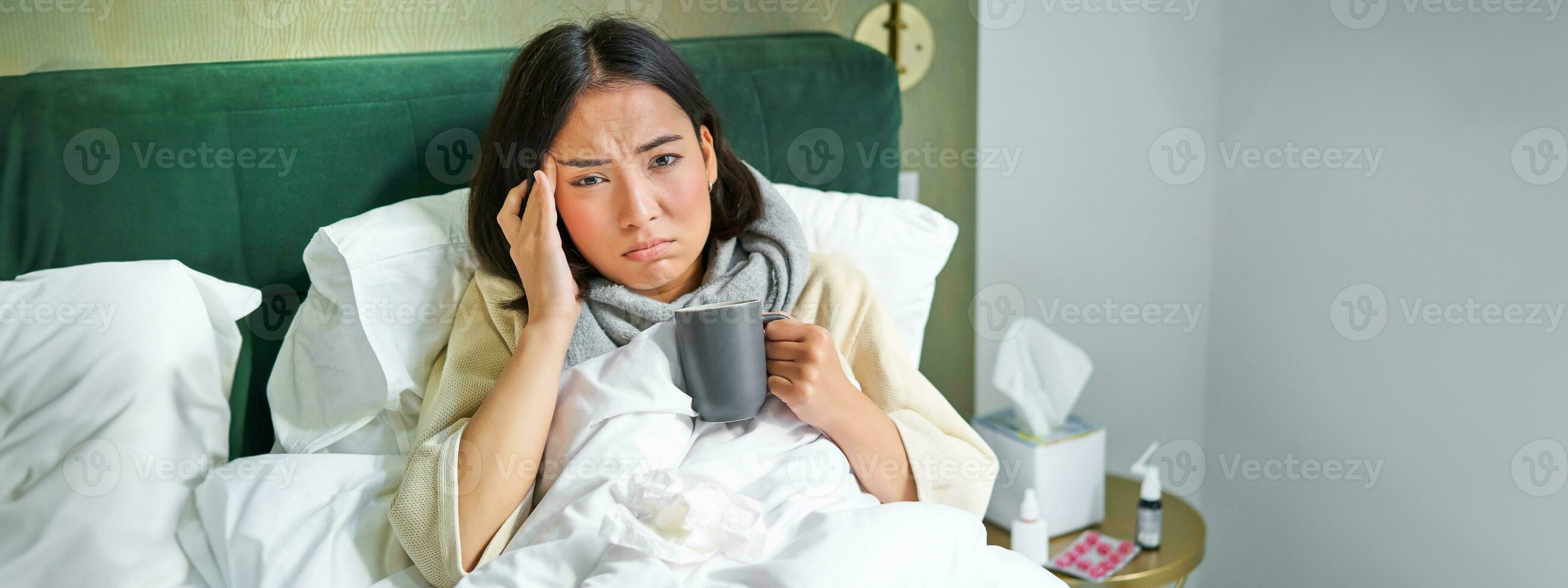 Portrait of asian woman with headache, catching col, staying on sick leave at home, lying in bed, drinking hot tea, having flu photo