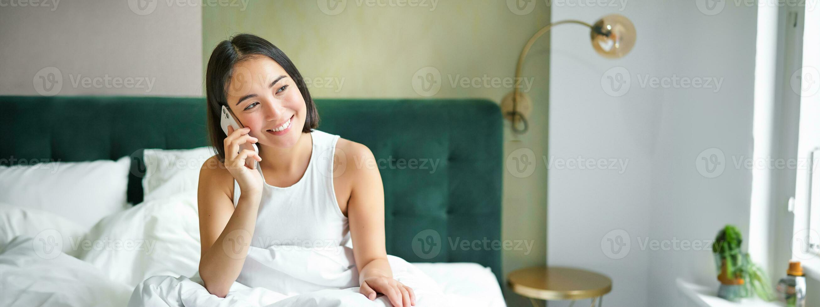 Smiling korean girl in bed, talks on mobile phone, making a phone call, lazy morning as asian woman orders delivery via smartphone photo