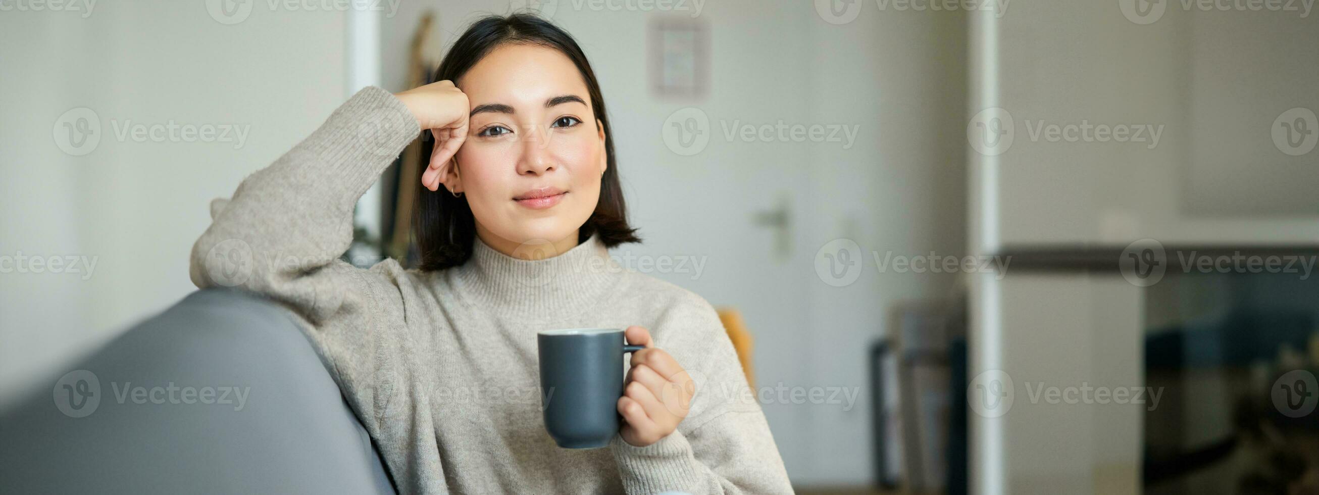 Smiling asian woman sitting on sofa with her mug, drinking coffee at home and relaxing after work, looking calm and cozy photo