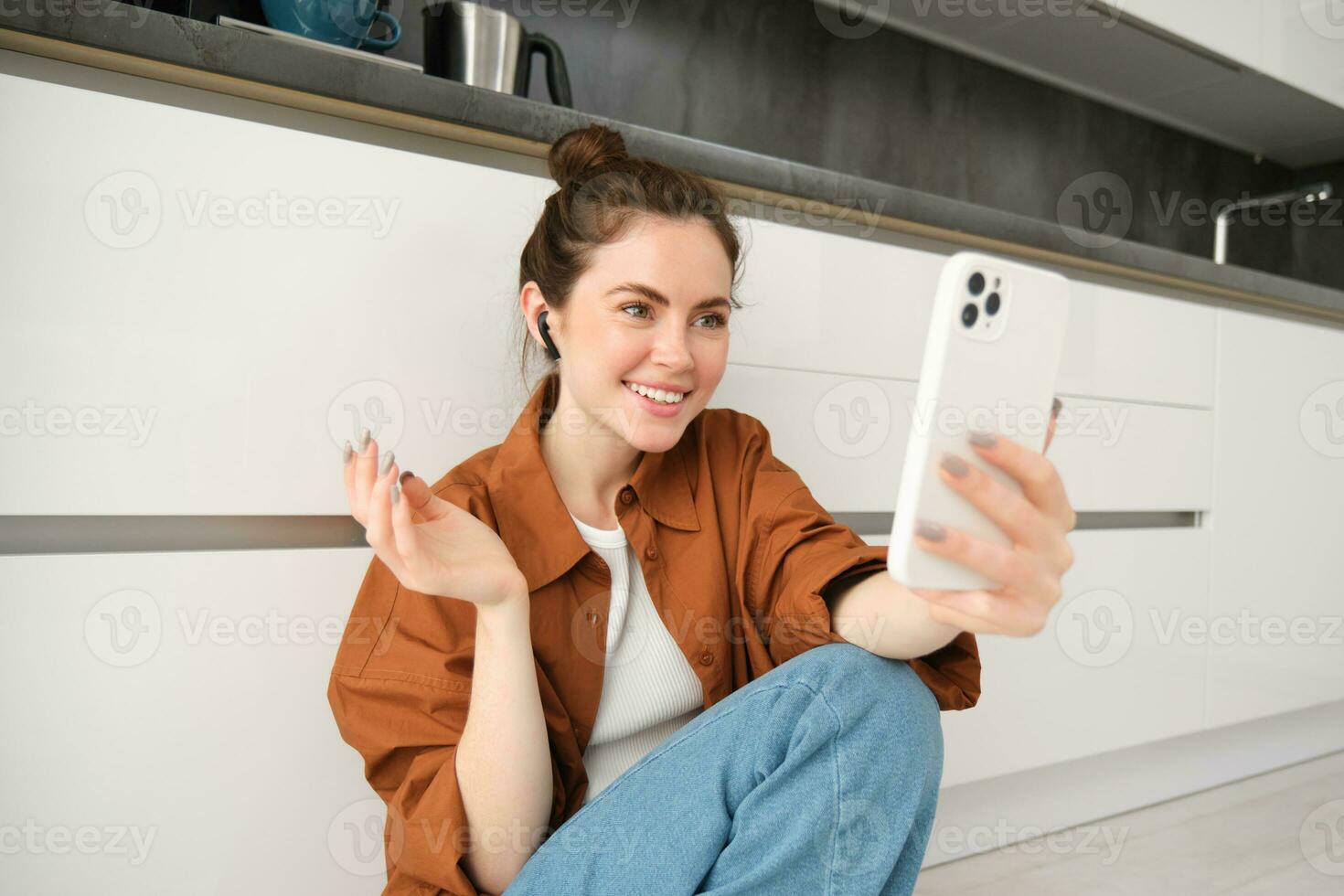 Friendly smiling woman talking on mobile phone app, using wireless earphones, looking at screen, online chatting, video calling friend, sitting on kitchen floor photo